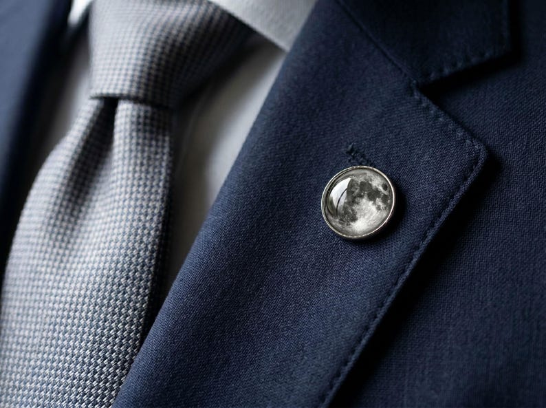 May include: A close-up of a navy blue suit jacket with a gray tie. A silver lapel pin with a detailed moon design is affixed to the jacket. The tie has a textured pattern.