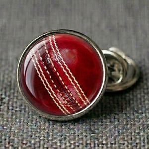 May include: A pair of silver cufflinks featuring a cricket ball design. The cufflinks have a round, polished silver frame. The cricket ball is red with white stitching, showcasing the details of the ball.