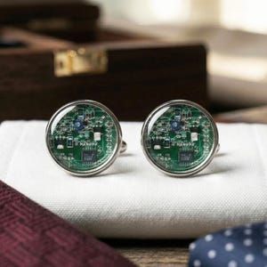 以下が含まれることがあります： 回路基板のデザインが施された、銀色のカフスボタンのペア。 カフスボタンは透明なドーム型の表面を持ち、回路基板の緑と銀色の詳細を拡大します。 白い折り畳まれた布の上に置かれ、背景にはバーガンディのネクタイと青い水玉模様のネクタイがあります。
