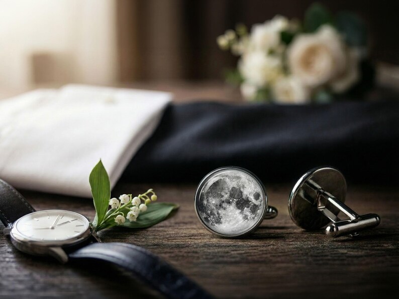May include: A pair of silver cufflinks with a detailed moon design, next to a silver wristwatch with a black leather band. A white dress shirt and a dark navy blue fabric are in the background, with a bouquet of white flowers.
