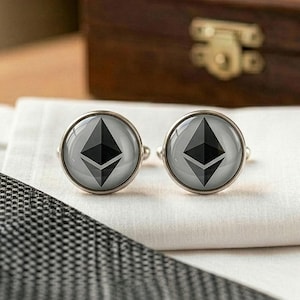 May include: A pair of silver cufflinks with a gray background and a black Ethereum symbol. The cufflinks are round and sit on a white cloth, with a patterned tie in the foreground and a wooden box in the background.