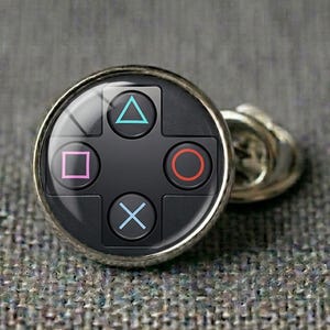 Op de afbeelding: Zilverkleurige manchetknop met een zwarte voorkant met de iconische PlayStation-controllerknopsymbolen: een blauwe driehoek, een roze vierkant, een rode cirkel en een blauwe X. De manchetknop is rond met een glanzende zilveren rand.