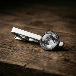 May include: A silver-toned tie clip with a circular, black and white image of the moon. The clip is rectangular with a rounded end and is set against a dark wood grain surface.