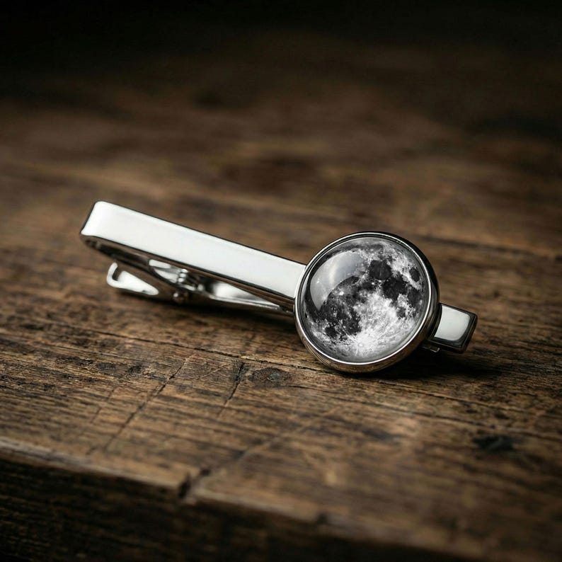 May include: A silver-toned tie clip with a circular, black and white image of the moon. The clip is rectangular with a rounded end and is set against a dark wood grain surface.
