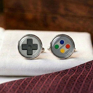 May include: A pair of silver cufflinks with a gaming theme. One cufflink features a directional pad design, while the other displays colored buttons. The cufflinks are set against a white background with a burgundy patterned tie.