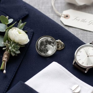 May include: Navy blue suit jacket with a white flower boutonniere, moon-themed cufflinks, a silver watch with a brown leather strap, and a tag that says "For the Groom."