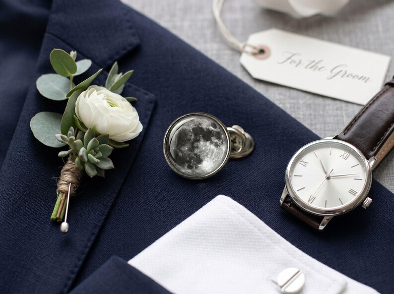 May include: Navy blue suit jacket with a white flower boutonniere, moon-themed cufflinks, a silver watch with a brown leather strap, and a tag that says "For the Groom."