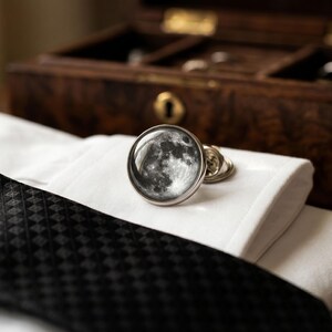 May include: A pair of silver cufflinks with a detailed moon phase design, resting on a white dress shirt cuff. A black patterned tie is visible, with a wooden jewelry box in the background.