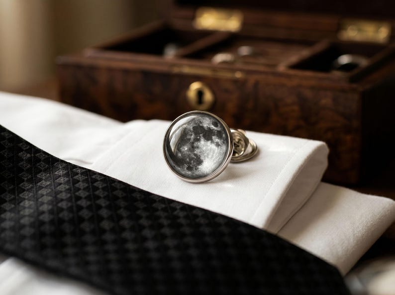 May include: A pair of silver cufflinks with a detailed moon phase design, resting on a white dress shirt cuff. A black patterned tie is visible, with a wooden jewelry box in the background.