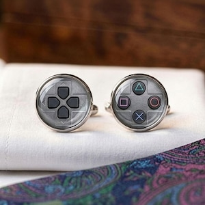 May include: A pair of silver cufflinks with a gaming controller design. One cufflink features a directional pad, while the other displays the action buttons. The cufflinks are set against a white background with a patterned fabric.
