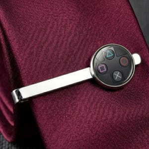 May include: Silver-toned tie clip featuring a black circular design with colorful symbols, resembling a video game controller. The tie clip is displayed on a burgundy tie, showcasing a sleek and modern accessory.
