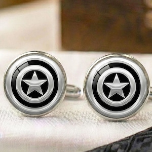 Może przedstawiać: Para okrągłych spinek do mankietów o monochromatycznym wzorze z centralnie umieszczoną pięcioramienną gwiazdą otoczoną koncentrycznymi pierścieniami. Metaliczne wykończenie tworzy odblaskową powierzchnię o kontrastujących czarnych i srebrnych odcieniach.