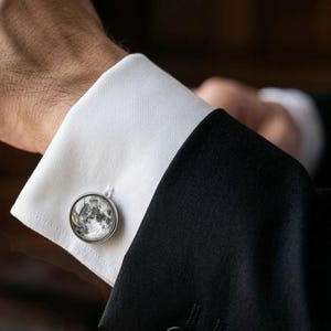 May include: A close-up of a silver cufflink with a detailed moon phase design. The cufflink is attached to a white dress shirt cuff, paired with a black suit jacket. The image highlights the elegant accessory.