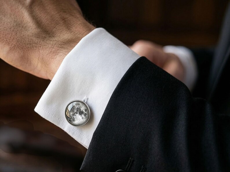May include: A close-up of a silver cufflink with a detailed moon phase design. The cufflink is attached to a white dress shirt cuff, paired with a black suit jacket. The image highlights the elegant accessory.