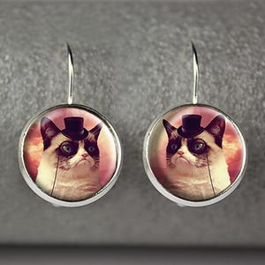 May include: A pair of silver-toned dangle earrings. Each earring features a round, glass-covered image of a cat wearing a top hat and monocle. The cat has a grumpy expression, with a white face and dark ears.