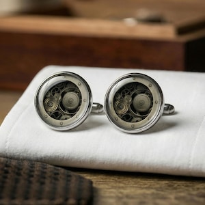 May include: Silver cufflinks with a steampunk design. Each cufflink has a clear, circular face displaying a detailed arrangement of bronze-coloured gears. The cufflinks are set against a white background, suggesting formal wear.
