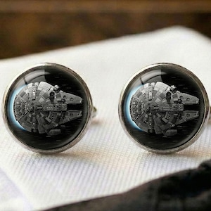 Op de afbeelding: Een paar ronde, zilverkleurige manchetknopen met een gedetailleerde zwart-witafbeelding van het Millennium Falcon-ruimteschip uit Star Wars. Het ronde ontwerp is gevat onder een heldere glazen koepel in een gepolijst metalen frame.