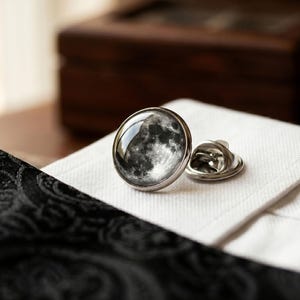May include: A silver-toned lapel pin with a detailed image of the moon. The pin is round with a domed glass or resin covering the lunar surface. It rests on a white, textured fabric, next to a black patterned fabric.