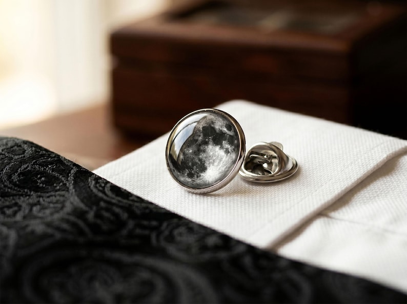 May include: A silver-toned lapel pin with a detailed image of the moon. The pin is round with a domed glass or resin covering the lunar surface. It rests on a white, textured fabric, next to a black patterned fabric.