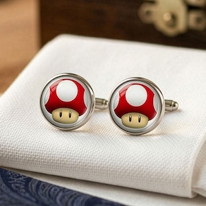 May include: A pair of silver cufflinks featuring a red and white mushroom design with a tan face. The cufflinks are set against a white cloth background, with a dark blue patterned fabric visible below.