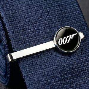 Op de afbeelding: Zilveren stropdasklem met een zwart cirkelvormig embleem met de witte tekst "007" en een pistoolsilhouet. De stropdasklem is geplaatst op een gestructureerde marineblauwe stropdas.