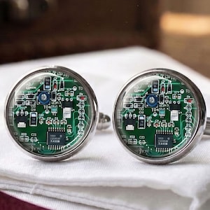 May include: A pair of round cufflinks featuring a green circuit board pattern under a clear glass dome. The electronic design includes visible microchips, resistors, and printed white text labels like TD4 and X4118 set in a polished silver-tone metal frame.