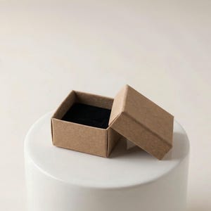 May include: An open, small, square, cardboard jewelry box with a black velvet interior. The box is light brown and sits on a white, cylindrical surface. The lid is slightly ajar, revealing the interior.