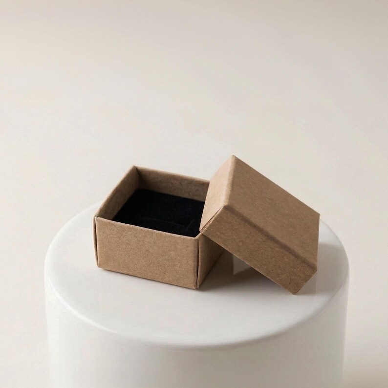 May include: An open, small, square, cardboard jewelry box with a black velvet interior. The box is light brown and sits on a white, cylindrical surface. The lid is slightly ajar, revealing the interior.