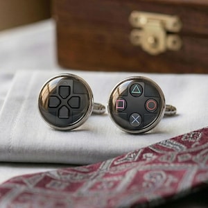 May include: A pair of silver cufflinks with a black face, each featuring a video game controller button design. One cufflink displays directional buttons, the other shows colored action buttons. The cufflinks rest on a white surface.