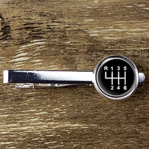 Può includere: Fermacravatta argentato con un design circolare nero con un motivo di cambio marcia bianco, inclusi i numeri R, 1, 3, 5, 2, 4 e 6. Su sfondo in legno.