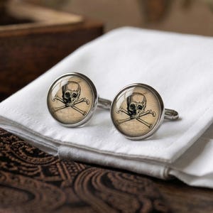 May include: A pair of silver cufflinks featuring a skull and crossbones design. The skull and bones are set against a tan background. The cufflinks are resting on a white folded cloth.