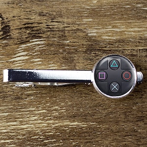 May include: A silver tie clip with a black circular button featuring a video game controller design. The button has a black background with a white X, a circle, a square, and a triangle.