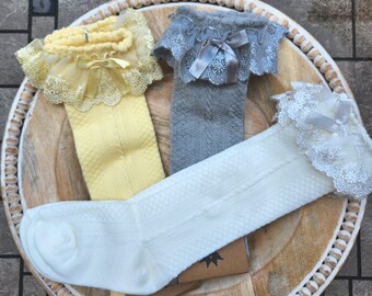 May include: Three pairs of knee-high socks in yellow, gray, and white. Each pair features a textured diamond knit pattern on the leg and a decorative lace ruffle cuff at the top, accented with a small matching satin ribbon bow at the center.