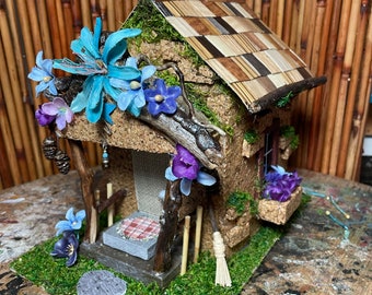 May include: A miniature fairy house made of cork with a moss-covered roof and a checkered roof. The house has a small porch with a checkered picnic blanket and a broom leaning against the wall. The house is decorated with blue and purple flowers.