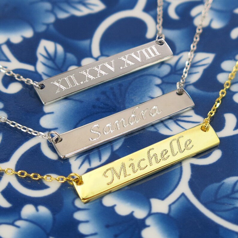 May include: Three silver bar necklaces with different engravings. The top necklace has Roman numerals XII.XXV.XVIII, the middle necklace has the name Sandra, and the bottom necklace has the name Michelle.