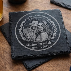 May include: Square black slate coaster with a laser-etched image of a couple, surrounded by a laurel wreath. The text "Wedding Anniversary", "Michael ♥ Jessica", and "June 18, 2025" are also etched on the coaster. The coaster is sitting on top of another coaster.