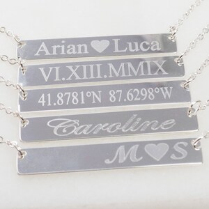 May include: Five silver bar necklaces with different engraved text. The top necklace says "Arian Luca" with a heart symbol between the names. The second necklace says "VI.XII.MMIX". The third necklace says "41.8781&deg;N 87.6298&deg;W". The fourth necklace says "Caroline". The bottom necklace says "M S" with a heart symbol between the letters.