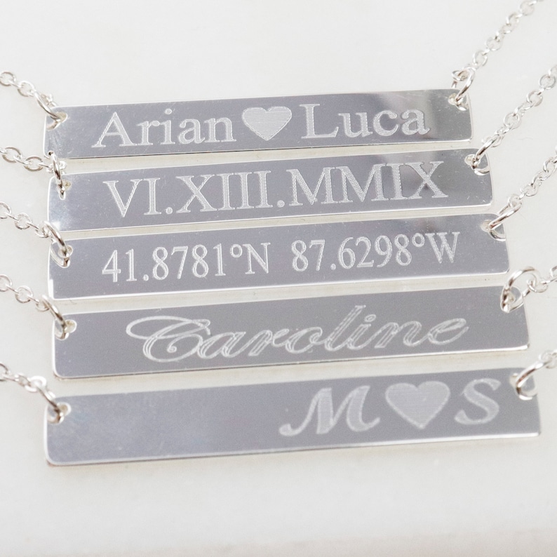 May include: Five silver bar necklaces with different engraved text. The top necklace says "Arian Luca" with a heart symbol between the names. The second necklace says "VI.XII.MMIX". The third necklace says "41.8781&deg;N 87.6298&deg;W". The fourth necklace says "Caroline". The bottom necklace says "M S" with a heart symbol between the letters.