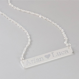 May include: A silver necklace with a bar pendant engraved with the names "Arian" and "Luca" separated by a heart symbol.