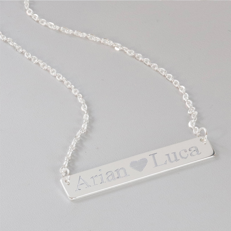 May include: A silver necklace with a bar pendant engraved with the names "Arian" and "Luca" separated by a heart symbol.