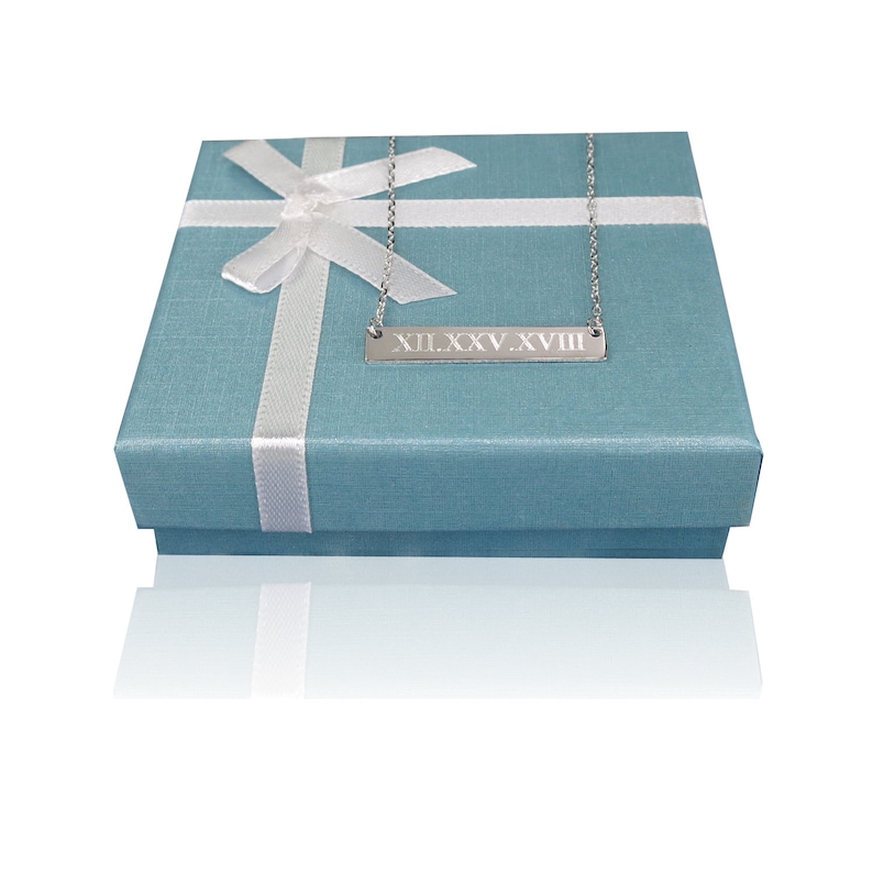 May include: A silver necklace with a bar pendant engraved with the Roman numerals "XII.XXV.XVIII" in a light blue gift box with a white ribbon.