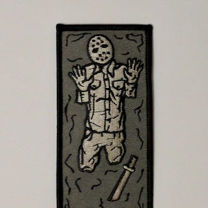 May include: A gray and black embroidered patch of a hockey mask-wearing figure in a white shirt and pants, with a knife, lying in a carbonite-like block.