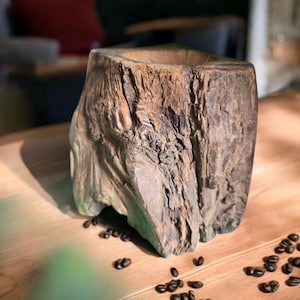 Puede incluir: Un jarrón de madera rústico y ahuecado con una superficie rugosa y texturizada. El jarrón es de color marrón natural y está sobre una mesa de madera, con granos de café esparcidos cerca. El fondo está borroso.