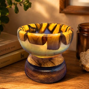 May include: A decorative bowl crafted from a combination of natural wood segments and translucent blue resin. The bowl features a wide, rounded upper section with visible wood grain and a solid, cylindrical wooden base with blue resin accents.