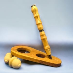 May include: A wooden pen with a natural wood finish and a silver band is displayed on a wooden stand. The stand has two holes, one for the pen and one for a small wooden object.