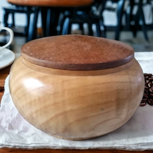 Könnte beinhalten: Ein Holzbehälter mit Deckel aus hellem braunem Holz. Der Behälter steht auf einem weißen Tuch mit Kaffebohnen, die um ihn herum verstreut sind.
