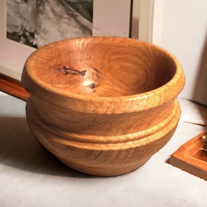 Puede incluir: Un cuenco de madera con un acabado de madera natural. El cuenco tiene una forma redondeada y un borde ligeramente acampanado. El cuenco está hecho de madera marrón claro con vetas marrones más oscuras.