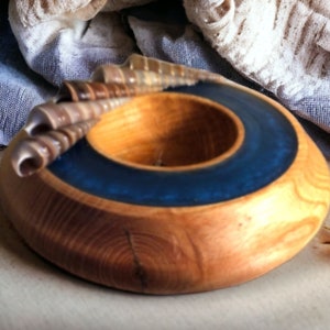 May include: A wooden bowl with a blue resin inlay. The bowl is round and has a natural wood finish. It is decorated with a cluster of seashells.
