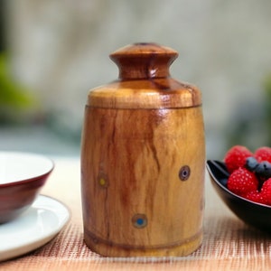 May include: A wooden jar with a lid and colorful dots on the side. The jar is sitting on a table with a bowl of fruit and a plate.