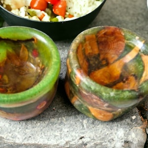 May include: Two small green and brown resin bowls with a mottled, wood-like pattern. The bowls are empty and sitting on a grey, textured surface.